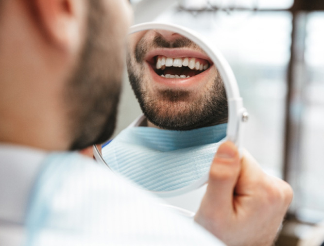 A young, bearded man admiring his smile with a hand mirror