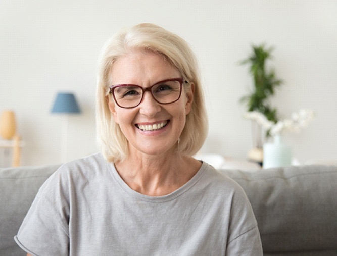 An older woman wearing glasses and a t-shirt and smiling because of her new and improved implant dentures